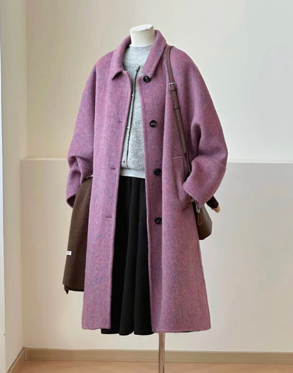 A lavender-purple wool-blend coat displayed on a mannequin, styled with a gray knit top, black skirt, brown shoulder bag, and dark brown lace-up boots on a floor stand.