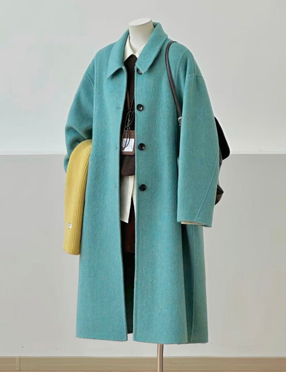 A long teal coat displayed on a mannequin stand, styled with a white shirt, dark tie, black bag, yellow clutch, and dark brown lace-up shoes on a light wood floor against a neutral wall.