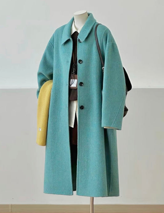 A long teal coat displayed on a mannequin stand, styled with a white shirt, dark tie, black bag, yellow clutch, and dark brown lace-up shoes on a light wood floor against a neutral wall.